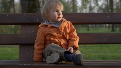 Portrait of little boy sitting on bench in park. Cute kid spending time outside.