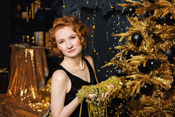 Model girl holding beads garlands on the background of a Christmas tree with gold decorations. Stylish women's, fashionable evening wear and accessories, makeup. Happy New Year.