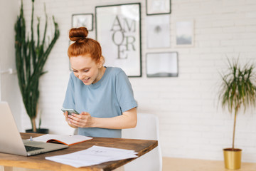 Portrait of happy cheerful redhead young business woman using mobile phone while sitting at the desk with laptop in light cozy room with modern interior at home office.