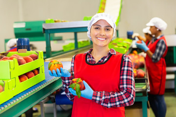 Smiling Latina woman worker standing in fruits industrial production facility demonstrating ripe...