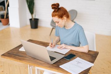 Creative redhead young woman designer is writing in documents, working on laptop while sitting at the desk in light cozy living room at home office. Concept of remote working.