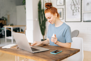 Cheerful young redhead woman is cleaning keyboard of laptop computer with sanitizer before starting work in light room at the home office. Concept of remote work or education from home 