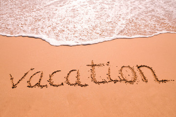 Word Vacation hand written in the sand with a sea wave. Close up sand texture on beach in summer.