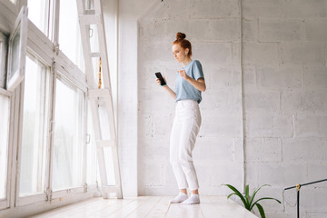 Fototapeta na wymiar Beautiful redhead young woman wearing wireless earphones listening music and dancing near big panoramic window. Pretty girl enjoying music on white background. Lady holding cell phone in hands.