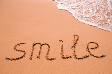 Word Smile hand written in the sand with a sea wave. Close up sand texture on beach in summer.