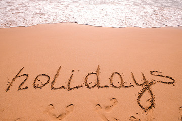 Word Holidays hand written in the sand with a sea wave. Close up sand texture on beach in summer.