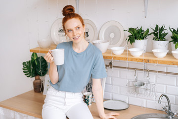 Beautiful redhead young woman holding cup of hot coffee while standing in kitchen room with white modern interior. Smiling business lady drinking tasty beverage from mug at home office in during break