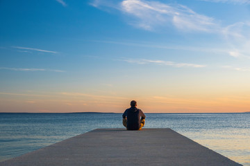 Man in hoodie and short cargo sitting on the edge of dock and meditating. Sea horizon, island od...