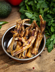 Dried chicken legs in the dog vessel among the greenery on the wooden board and some vegetables on the background. Chewing treats for domestic dogs. Natural dental treats for promotion and pampering.