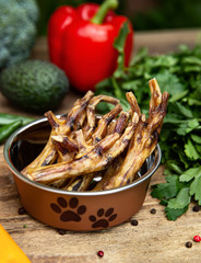 Dried chicken legs in the dog vessel among the greenery on the wooden board and some vegetables on the background. Chewing treats for domestic dogs. Natural dental treats for promotion and pampering.