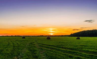 Obraz premium Scenic view at beautiful sunset in green shiny field with hay stacks, bright cloudy sky, country road and golden sun rays with glow, summer valley landscape