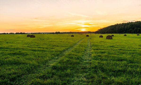 Scenic view at beautiful sunset in green shiny field with hay stacks, bright cloudy sky, country road and golden sun rays with glow, summer valley landscape © Yaroslav