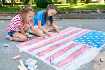 Two cute friends girls drawing American flag with colored chalks on the sidewalk near the house on sunny summer day. Kids painting outside. Creative development of children. Patriotic day concept