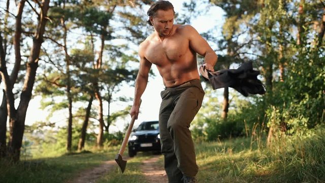 Shirtless man with muscular type of body cutting wood by axe in the forest near his car.