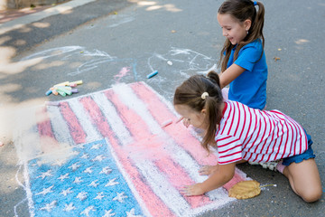Two cute friends girls drawing American flag with colored chalks on the sidewalk near the house on sunny summer day. Kids painting outside. Creative development of children. Patriotic day concept