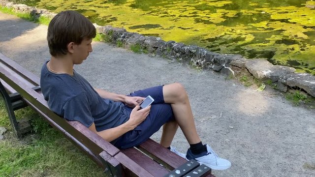 The young blonde guy is sitting on the bench and surfing the mobile phone. Man is sitting outside on the bench near the lake in summer. Look into mobile phone. Guy messaging in mobile. Tapping mobile