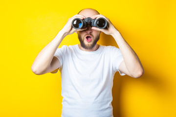 Surprised shocked bearded young man looks through binoculars on a yellow background