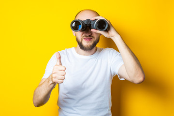 Smiling bald young man with a beard looks through binoculars and shows a thumb up on a yellow background