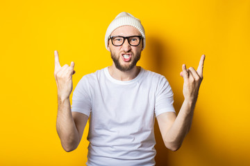 Bearded young man with glasses shows a rocker goat gesture on a yellow background.