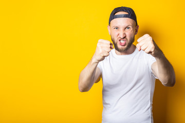 Young man with a beard in a baseball cap with clenched fists wants to punch, trains on a yellow background.