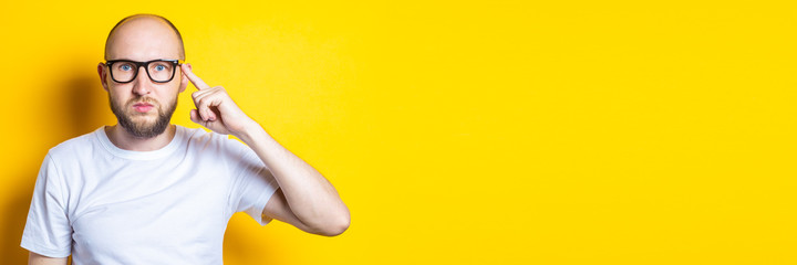Serious bearded bald young guy in glasses holds a finger to his temple on a yellow background....