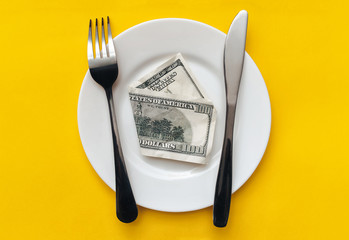 A hundred-dollar bill lies on a white plate between a fork and a knife on a yellow tablecloth top...