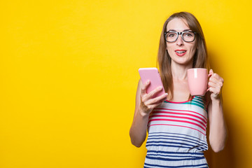 Young woman in striped dress drinks coffee with phone on yellow background.