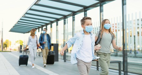 Cute small Caucasian happy kids, brother and sister in medical masks running at train station. Parents walking on blurred background and carrying suitcases on wheels. Trip concept in pandemic.