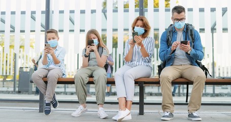 Social distance at bus stop among family members. Caucasian parents and kids in medical masks sitting on bench and using or playing on mobile phones. Tapping and scrolling on smarphones. Pandemic trip