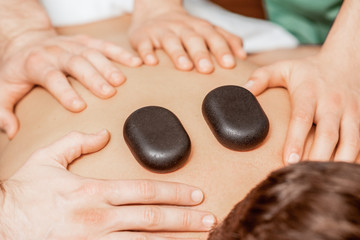 Hands of massage therapists doing back massage while hot stones on back of man close up in spa.