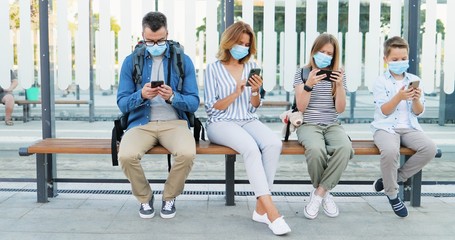Fototapeta na wymiar Social distance at bus stop among family members. Caucasian parents and kids in medical masks sitting on bench and using or playing on mobile phones. Tapping and scrolling on smarphones. Pandemic trip