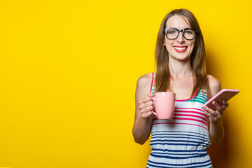 Beautiful happy smiling young girl holding a cup with coffee and a phone on a yellow background