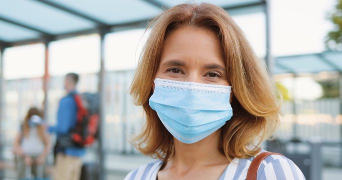 Portrait of beautiful Caucasian blonde woman in medical mask looking at camera and standing at bus stop or train station. Husband with kids and suitcases waiting for transport on blurred background.