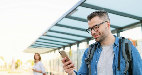 Caucasian handsome young man in glasses and with backpack standing at bus stop and tapping on mobile phone. Male in eyeglasses texting message on cellphone at train station.