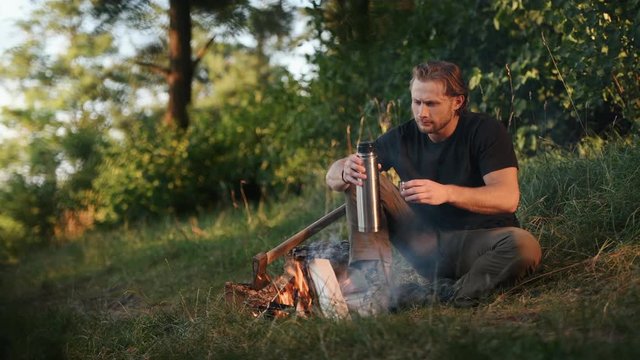 Man in black shirt sits by campfire in the forest at his weekend time and enjoying drink.