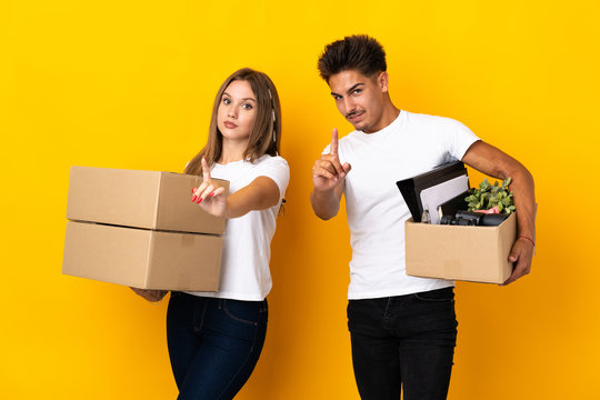 Teenager Couple Moving In New Home Among Boxes Isolated On Blue Background Showing And Lifting A Finger