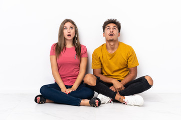 Young couple sitting on the floor isolated on white background looking up and with surprised expression