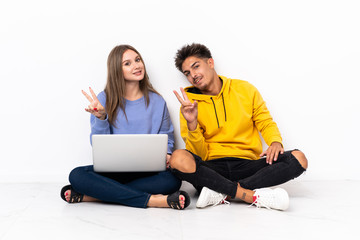 Young couple with a laptop sitting on the floor isolated on white background smiling and showing victory sign