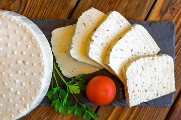 View of cheese head and cheese slicing on a black slate board served with tomatoes and fresh greens. Homemade cheese. Food concept. Selective focus.