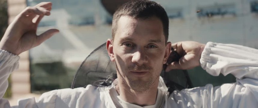 Portrait of a young beekeeper put on protective clothing veil or net for protection smile before work at beekeeping apiary with Apis mellifera carnica flying to wooden hives slowmotion close up