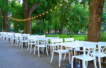 open air street cafe outdoor - white chairs and tables, decorated with illumination, the city Park and walking people