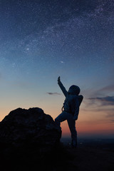 Space traveler standing on rocky mountain and pointing at stars. Silhouette astronaut wearing white space suit while looking at fantastic starry sky. Concept of galaxy, human in space and milky way.