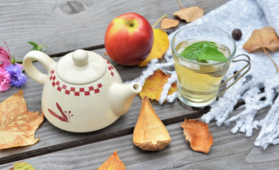 cup of tea  on a wooden table in garden with teapot  among autumnal  leaf and red apple on wool scarf