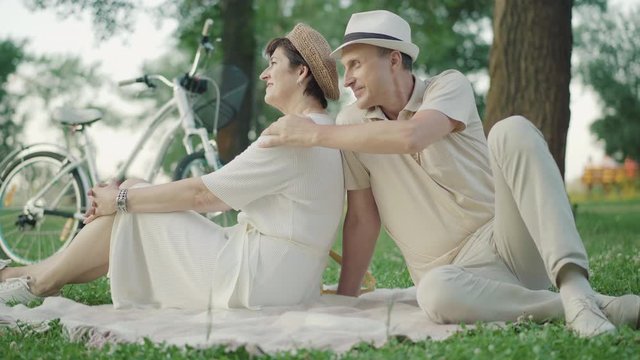 Elegant mid-adult Caucasian couple sitting on blanket in summer park and chatting. Portrait of happy positive man and woman enjoying sunset outdoors. Date of cheerful loving family.