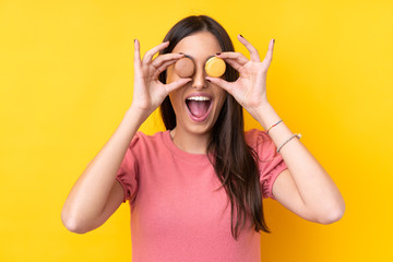Young brunette woman over isolated yellow background holding colorful French macarons with surprised expression