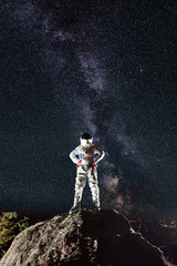 Astronaut standing on top of mountain with mesmerizing night sky with stars and Milky way on background. Space traveler in space suit keeping hands on waist. Concept of cosmonautics and space travel.