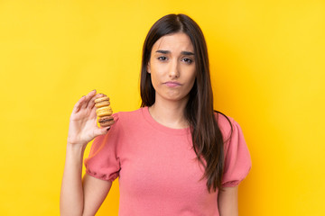 Young brunette woman over isolated yellow background holding colorful French macarons and with sad expression