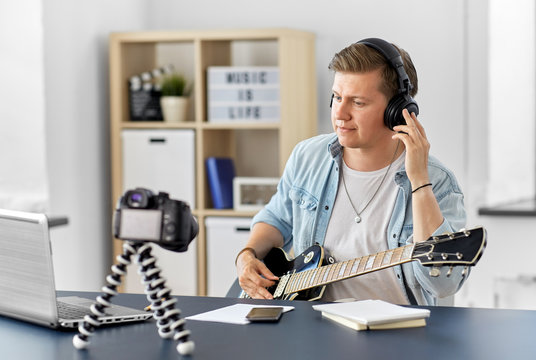 video blogging, music and people concept - young man or musician in headphones with camera videoblogging and playing guitar sitting at table at home