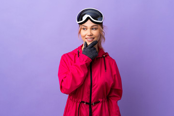 Skier teenager girl with snowboarding glasses over isolated purple background looking to the side and smiling