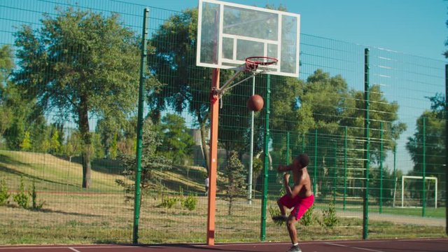 Shirtless muscular build african basketball player recieving pass from teammate, driving to hoop, jumping in the air and performing slam dunk with one hand while playing streetball game at urban court